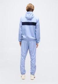 Άτομο που φοράει ανοιχτό μπλε φόρμα με σκούρα μπλε ρίγα στην πλάτη του μπουφάν, με κουκούλα, στέκεται γυρισμένο προς τα πίσω σε λευκό φόντο.