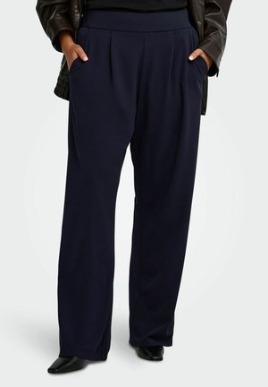 Personne portant un pantalon ample bleu marine taille haute avec des plis et des chaussures noires pointues, les mains dans les poches, une veste sombre visible en haut.