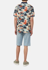 Homme portant une chemise à manches courtes avec un imprimé tropical, un short en denim clair jusqu'aux genoux et des baskets beiges, se tenant de dos.