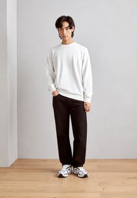 Ung mand iført en hvid langærmet skjorte, sorte bukser og hvide sneakers, der står på et trægulv foran en ensfarvet lys grå væg.