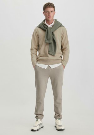 Beige Sweatshirt mit einem grünen Pullover, der um den Hals drapiert ist, kombiniert mit hellgrauen Sweatpants und weißen Turnschuhen mit grauen Akzenten.