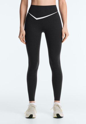 Femeie purtând colanți negri cu talie înaltă și model alb chevron în talie și pantofi sport bej, stând drept.