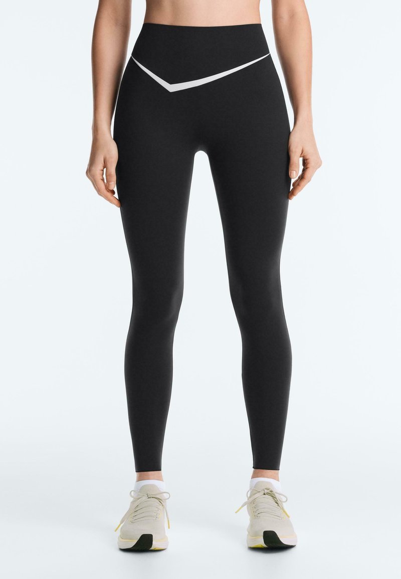 Femme portant des leggings noirs taille haute avec un motif chevron blanc à la taille et des chaussures de sport beiges, debout droite.