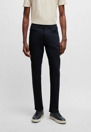 Pantalon marine slim fit en mélange de coton avec une texture lisse, doté d'une fermeture éclair, de passants de ceinture et de poches latérales, porté avec un t-shirt de couleur claire.