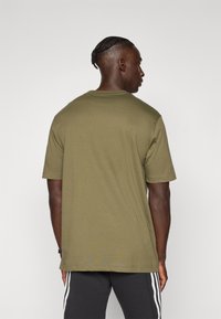 Mand med fletning iført en olivengrøn t-shirt og sorte bukser med hvide striber, der står vendt væk mod en ensfarvet baggrund.