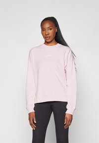Sudadera rosa claro con cuello redondo y mangas largas. Presenta un pequeño logo blanco en el pecho. Tejido suave con un corte relajado. Combinada con leggings negros.