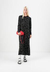 Svart langermet kjole med hvitt blomsterdesign, parret med en rød håndveske og svarte sandaler over hvite sokker. Enkel, løs design.