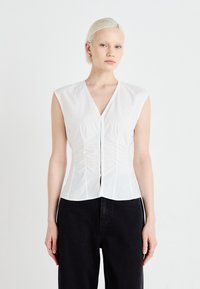 Blouse sans manches blanche avec un col en V, fronces sur le devant et coupe ajustée, fabriquée en tissu lisse. Associée à un pantalon noir à jambes larges.