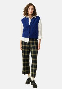 Blå stickad väst med diamantmönster, vit skjorta under, rutiga byxor i gröna och gula toner, svarta skor, gräddfärgade strumpor.