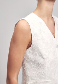 White sleeveless dress featuring floral embossed texture, v-neckline, and a subtle button detail, highlighting a clean, structured design.