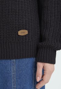 Mørk strikket genser med ribbestrikket kant, med et lærmerke merket "oxmo." Bært med blå denim, som viser et teksturert mønster.