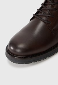 Brązowy skórzany boot z zaokrąglonym czubkiem, teksturowaną powierzchnią i kontrastującą gumową podeszwą o strukturze. Posiada sznurowadła oraz detale szycia.