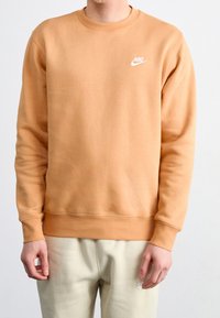 Man som bär en tanfärgad Nike-tröja och ljusbeige sweatpants, stående mot en enkel vit bakgrund.