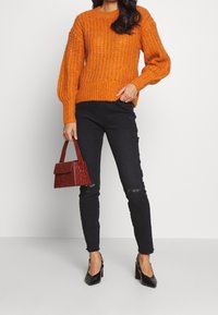Pull tricoté orange avec texture côtelée, jean noir déchiré et petit sac à main rouge embossé crocodile. Chaussures à talons noires.