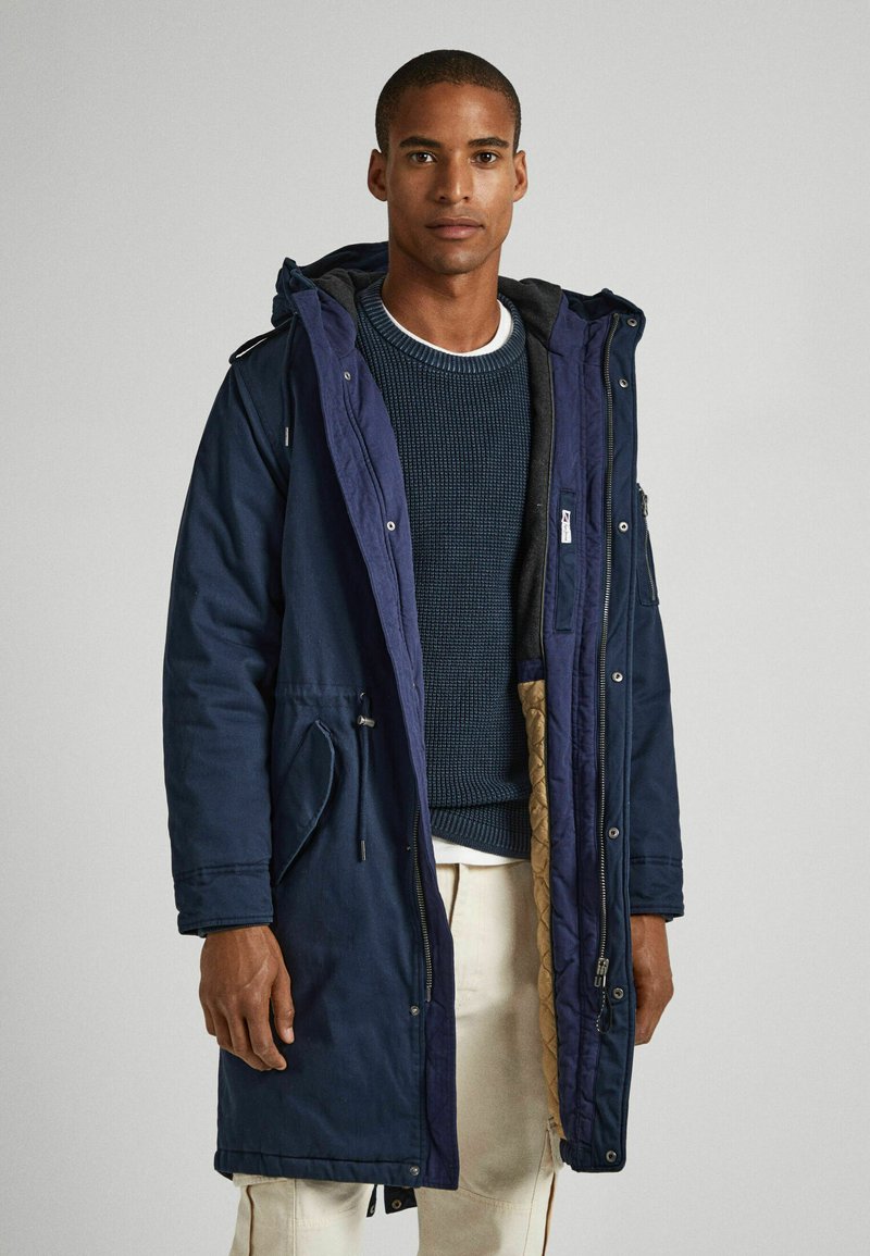 Parka della marina con cappuccio, vita regolabile con cordino, zip frontale e tasche laterali. Foderato con un materiale imbottito di colore beige. Pullover in maglia strutturata sotto.