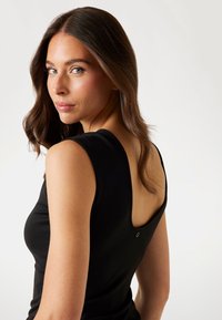 Černý top bez rukávů s nízkým zadním designem, hladkou texturou a malým logem blízko zapínání. Modelka předvádí oděv ze strany.