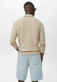 Suéter de punto beige con cuello de polo, mangas largas y patrón texturizado, combinado con pantalones cortos de denim azul claro y gafas de sol.