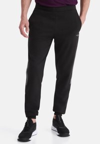 Schwarze Sweatpants aus weichem Stoff, mit schmalem Schnitt, seitlichen Streifen und einem elastischen Bund. Kombiniert mit schwarzen Sportschuhen.