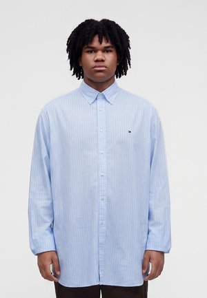 Jeune homme aux cheveux bouclés portant une chemise bleu clair à manches longues avec de fines rayures blanches, debout devant un fond uni.