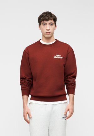 Noormees, kes kannab bordo värvi New Balance'i dressipluusi ja valgeid pükse, seisab ühevärvilise heledahalli tausta ees.