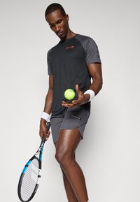 T-shirt de sport noir évacuant l'humidité avec des accents gris, associé à un short de sport gris. Tient une raquette de tennis et une balle de tennis vert fluo.
