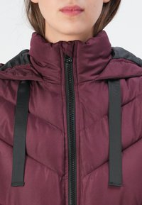 Veste en duvet bordeaux avec un col montant, des accents noirs, des coutures en zigzag et une fermeture à glissière sur le devant. Dispose de cordons noirs pour l'ajustement.
