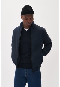 Jūros pajėgų bombonešio striukė su aukštu apykakle ir užtrauktuku, derinama su tamsiu megztiniu. Modelis dėvi juodą megztą beanie skrybėlę. Paprastas, elegantiškas dizainas.