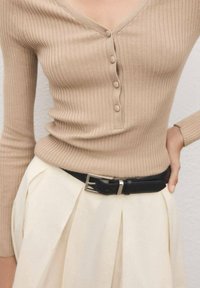 Top bej, cu dungi, cu mânecă lungă și nasturi, băgat în pantaloni crem cu talie înaltă, asigurat cu o curea neagră având cataramă argintie.