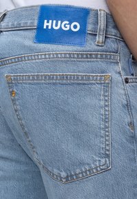 Πίσω μέρος ανοινομπλέ τζιν με ένα μπλε ορθογώνιο ετικέτα ραμμένη πάνω από την δεξιά τσέπη με την επιγραφή "HUGO" με λευκά γράμματα.