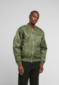 Giubbotto bomber verde oliva in nylon lucido, con chiusura frontale a zip, polsini a coste e logo ricamato sul lato sinistro del petto.
