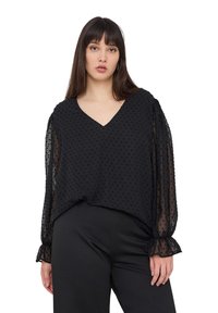 Blusa nera con scollo a V, caratterizzata da pois texturizzati e maniche puff trasparenti. Il tessuto ha un aspetto morbido e fluido. Indossata con pantaloni neri.
