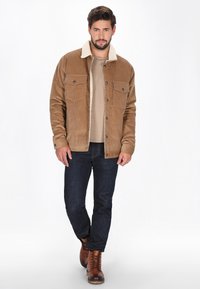Braune Cordjacke mit einem beigefarbenen Schafsfellkragen, Knopfverschluss vorne, zwei Brusttaschen, kombiniert mit dunklen Jeans und braunen Schnürstiefeln.