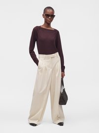 Personne portant un haut à manches longues foncé, un pantalon large de couleur crème, des chaussures noires, des lunettes de soleil, et tenant un petit sac à main noir.