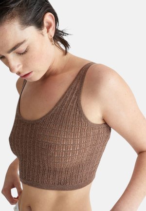 Giovane donna con capelli corti e scuri che indossa un top corto marrone texturizzato senza maniche su uno sfondo chiaro e uniforme.