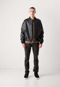 Zwart leren bomberjack met een doorgestikte voorkant, geribde manchetten en een ritssluiting, over een bruine shirt en grijze spijkerbroek. Zwarte sneakers.