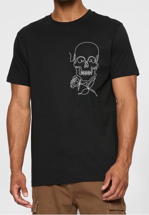 Mand iført sort T-shirt med hvidt enkeltlinjet motiv af kranium og rose på venstre bryst, kombineret med brune cargobukser.