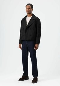 Zwarte dubbelgeknoopte peacoat met brede revers, gestructureerde stof, gecombineerd met donkere jeans en bruine schoenen. Model staat tegen een witte achtergrond.