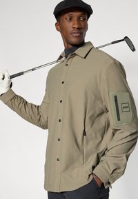 Πράσινο ελιάς τζάκετ γκολφ με υφασμάτινη υφή, μπροστινά κουμπιά και τσέπη με φερμουάρ που διαθέτει λογότυπο στο μανίκι. Κομψός, λειτουργικός σχεδιασμός.