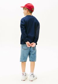 Marineblaues Langarmshirt, Jeansshorts und weiße Turnschuhe. Roter Hut des Kindes, Hände hinter dem Rücken verschränkt, vor einem schlichten Hintergrund stehend.