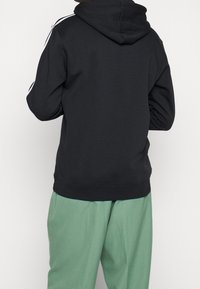 Schwarzer Hoodie mit einer frontalen Kängurutasche, verstellbarem Kordelzug an der Kapuze und weißen Streifenakzenten an den Schultern. Kombiniert mit grünen Hosen.