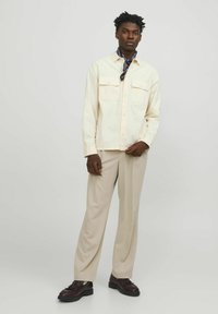 Chemise button-up crème clair avec deux poches poitrine, associée à un pantalon beige et des chaussures à lacets marron foncé. Tissu doux et coupe décontractée.