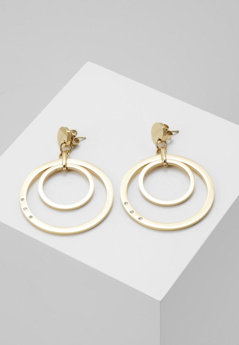 Paire de boucles d'oreilles créoles circulaires dorées avec des anneaux intérieurs plus petits et de petits détails en cristal, placées sur une surface blanche inclinée.