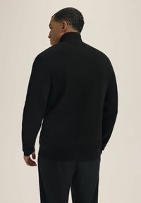 Czarny sweter z wysokim kołnierzem, o teksturowanej tkaninie i luźnym kroju. Widoczne detale szwów na ramionach i plecach.