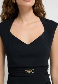 Musta mekko, jossa on v-pääntie ja hihattomat hihat, joissa on kultainen sisustus vyötäröllä. Sujuva kankaan rakenne sekä muotoillut yksityiskohdat.