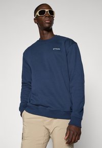 Man in beige broek, marineblauwe Columbia sweatshirt en grote crèmewitte zonnebril, geposeerd tegen een lichte achtergrond.