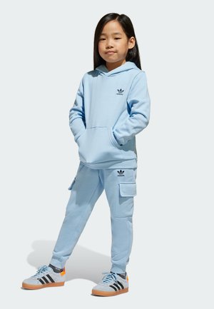 Felpa Adidas di colore blu chiaro con tasca a canguro, abbinata a pantaloni jogger coordinati con tasche cargo. L'outfit è completato da scarpe da ginnastica grigie e nere.