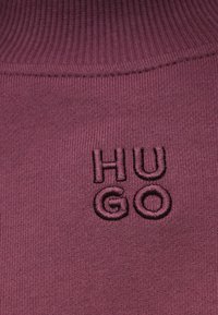 Burgunder farget strikkestoff med en ribbet tekstur og en brodert logo "HUGO" i en mørkere nyanse, hvor "H" og "G" skiller seg ut i størrelse.