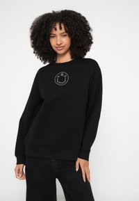 Μαύρο φούτερ με στρογγυλή λαιμόκοψη, με κυκλικό γραφικό ενός χαμογελαστού προσώπου και αρχικά της μάρκας σε ασημένιες στρας στο στήθος.