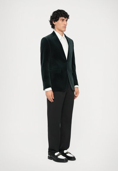 Pánský tmavě zelený sametový blazer, bílá košile, černé kalhoty a černé mokasíny s bílými akcenty. Blazer má dvě přední kapsy a slim fit střih.