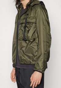 Person klädd i en olivgrön Nike-hoodie med svart dragkedja, framficka och svarta byxor, som står åt sidan mot en neutral bakgrund.